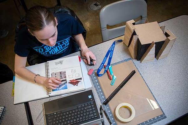 A student sits at a table, wearing a blue Bailey College of Technology T-shirt. A notebook is on the table and the student looks at a laptop. Cardboard, a ruler, and tape are to the left.  